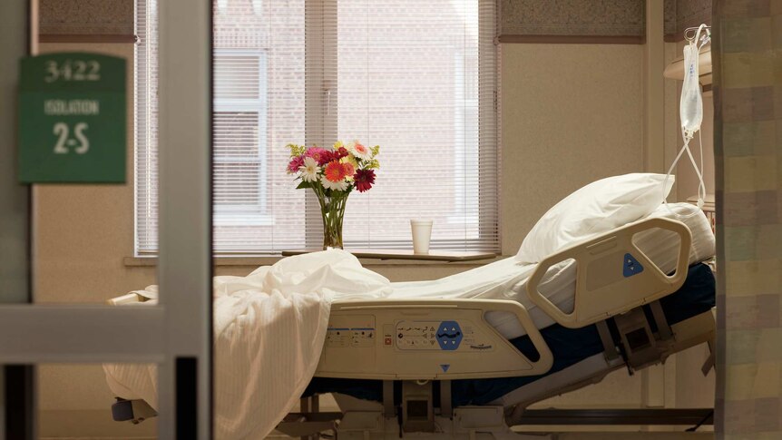 A vase of flowers on a window sill overlooks an empty hospital bed, lit with warm lighting.
