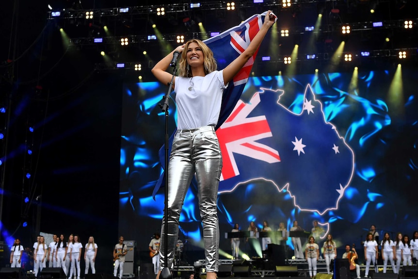 Delta Goodrem holds an Australian flag above her as a choir sings behind her on stage at a bushfire relief concert