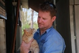 A man holds a ginger cat, which is staring up at him.