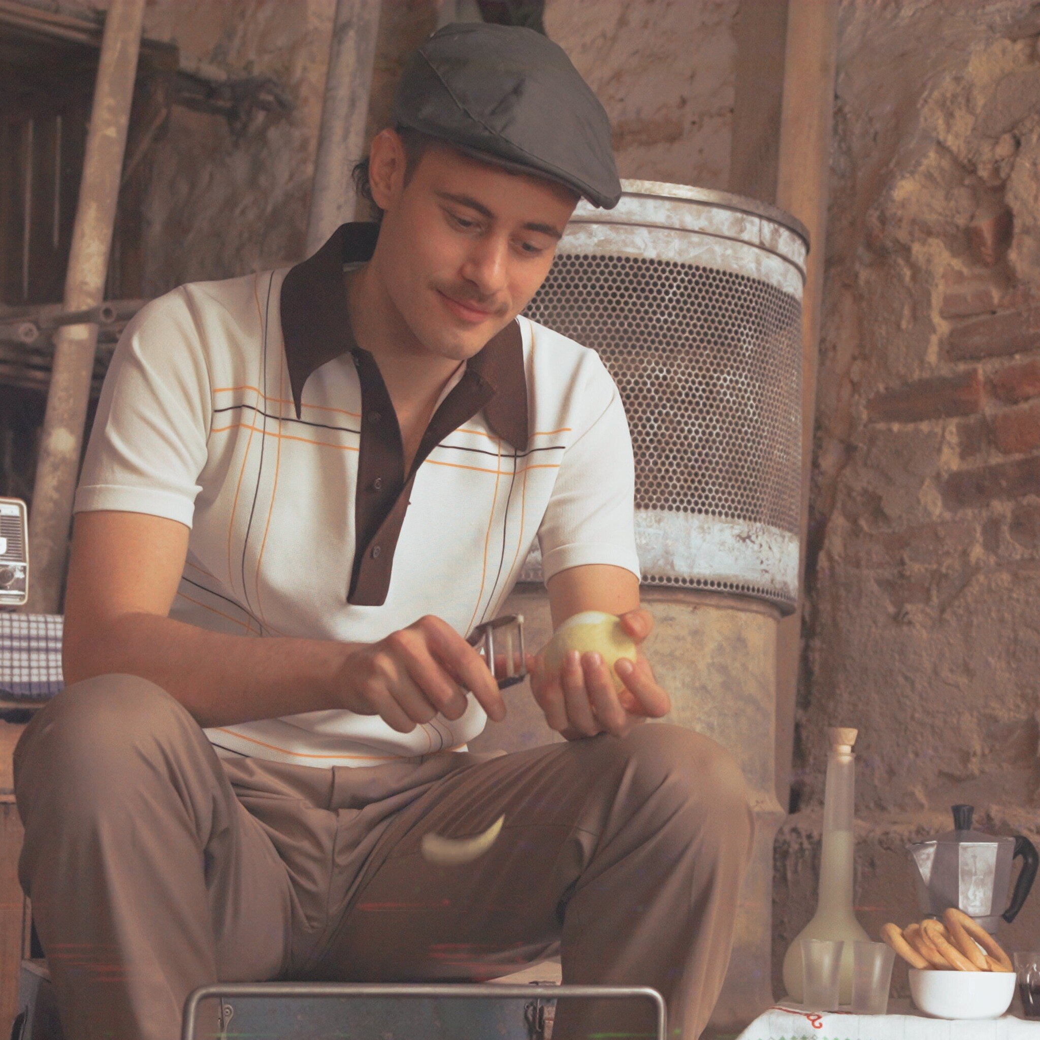 Un joven de piel clara y bigote pela un limón frente a una pared de ladrillos. 