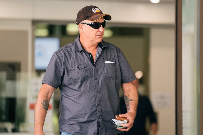 A man wearing a blue shirt, sunglasses and black cap walks outside a building carrying a wallet and other items.