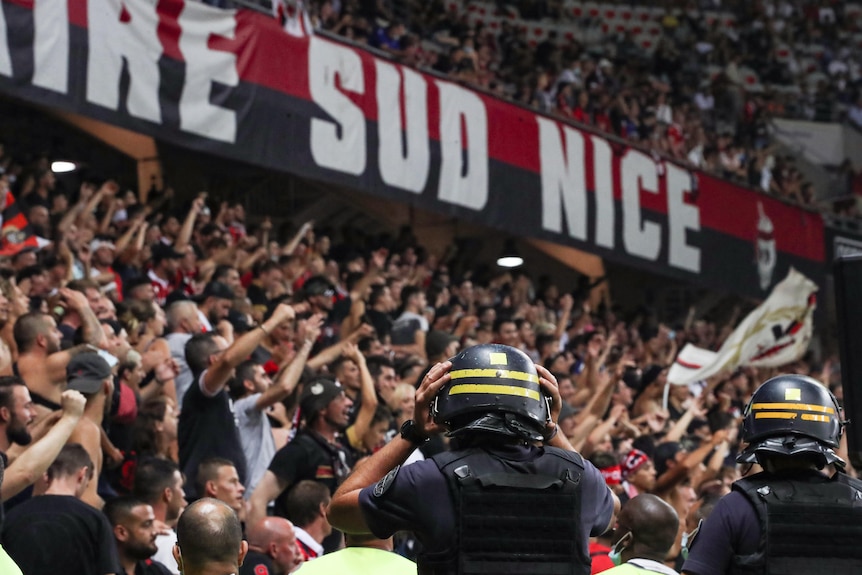 Des policiers blindés lourds en tenue animée regardent des fans de football scandant un stand