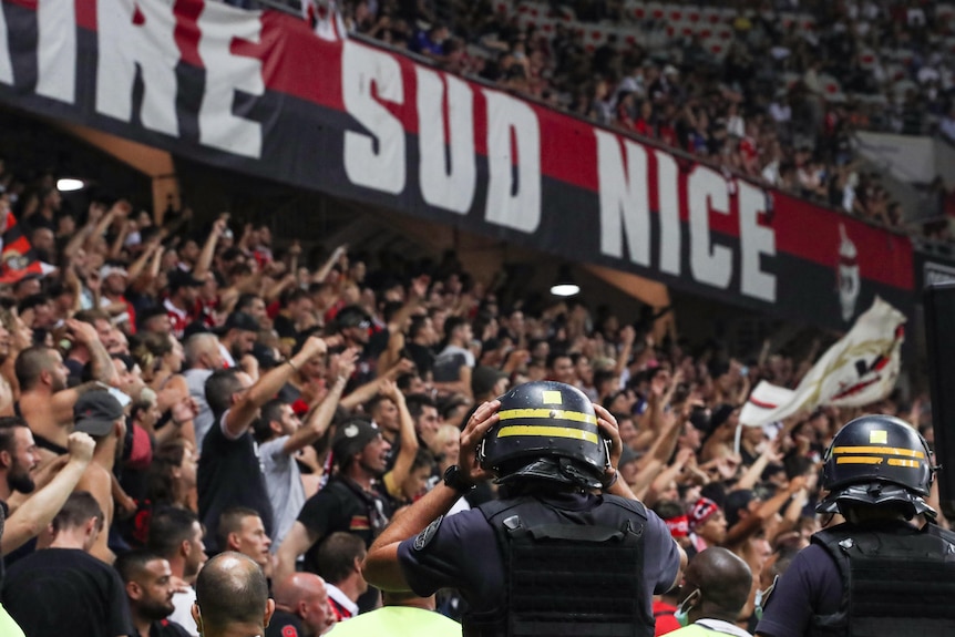 Heavily armoured police in riot gear watch chanting football fans in a stand