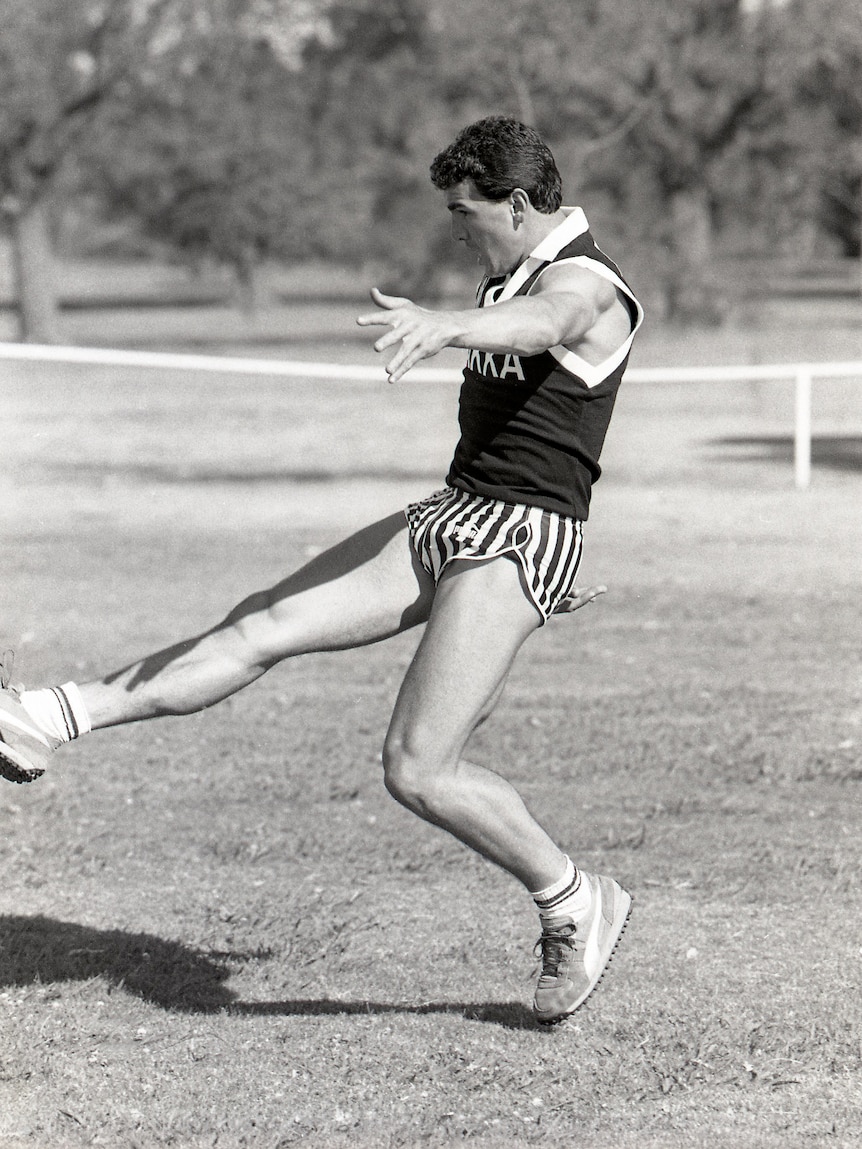 A black and white photo of Darren Millane in Collingwood training gear kicking a football.