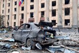 A burned out car is surrounded by debris in front of a damaged building