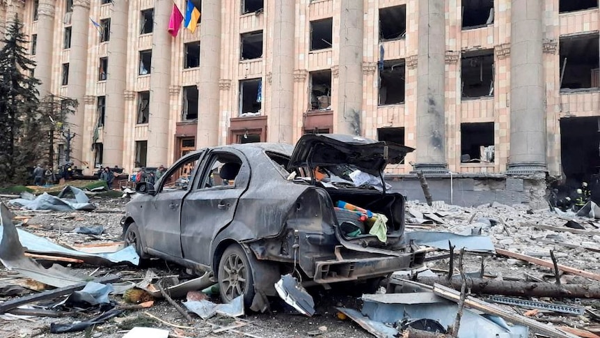 A burned out car is surrounded by debris in front of a damaged building