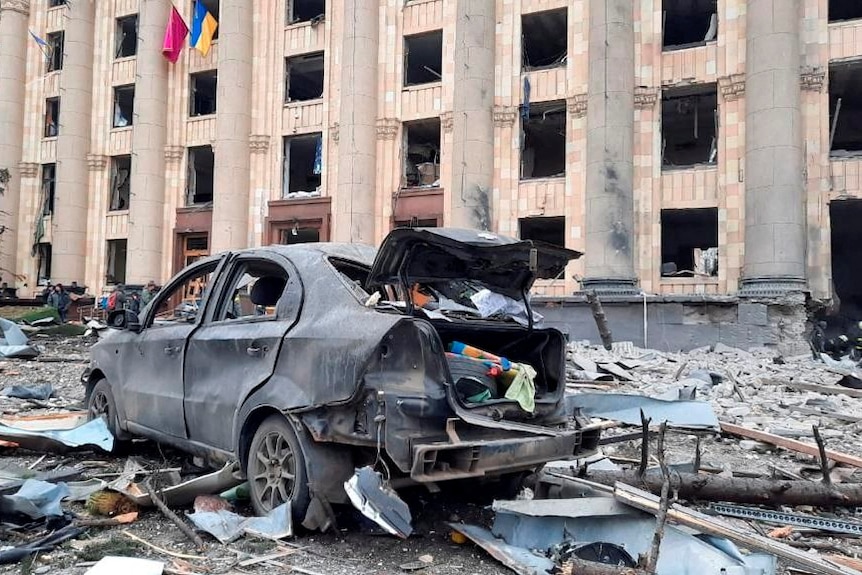 A burned out car is surrounded by debris in front of a damaged building