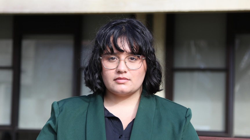 A head shot of a serious but determined looking young woman, short black bob, green jacket, round glasses, black top.