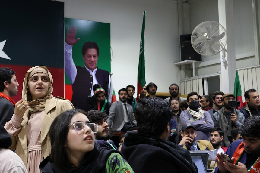 A crowd of people watch a TV screen with a large poster of Imran Khan in the background.