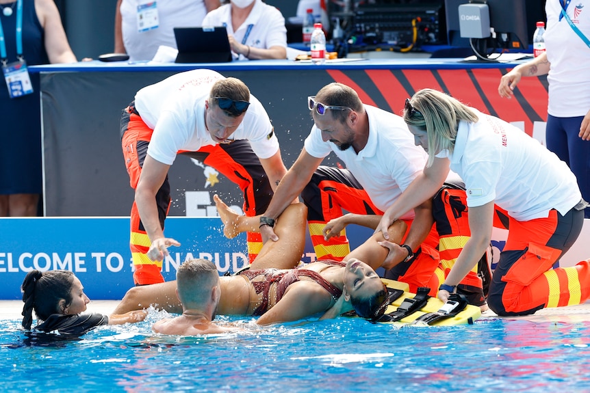 An unconscious woman is pulled from a swimming pool by three people and lifted onto a stretcher.