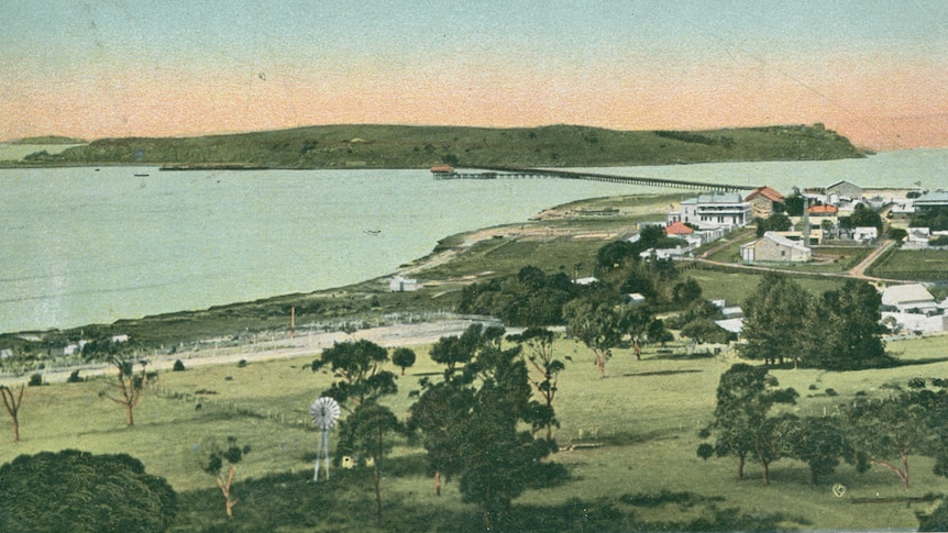 A historic coloured postcard showing a small town and island linked by a bridge