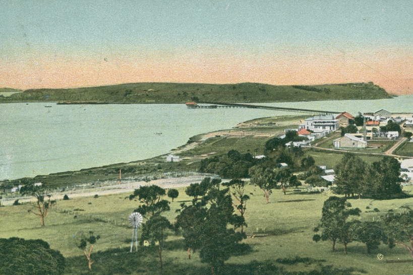 A historic coloured postcard showing a small town and island linked by a bridge