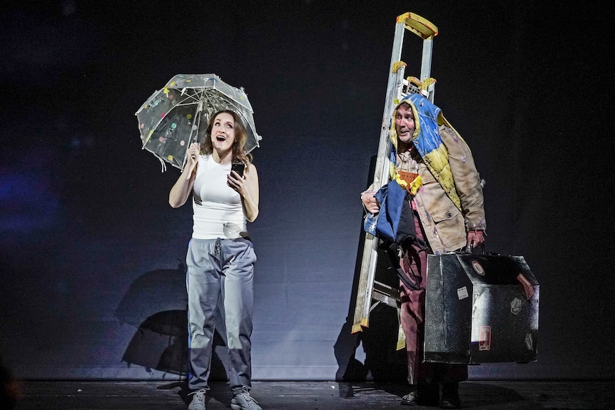 A white female opera singer holds an umbrella onstage beside a white male opera singer in painting clothes, singing.