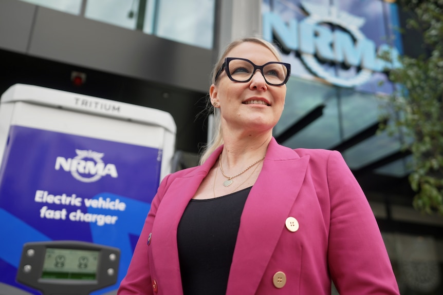 A woman wearing large black rimmed glasses and a pink blazer stands in front of an NRMA electric vehicle fast charging station.