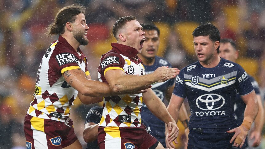 A Brisbane Broncos player stands and yells in celebration with a teammate as rain falls in a game against the Cowboys.