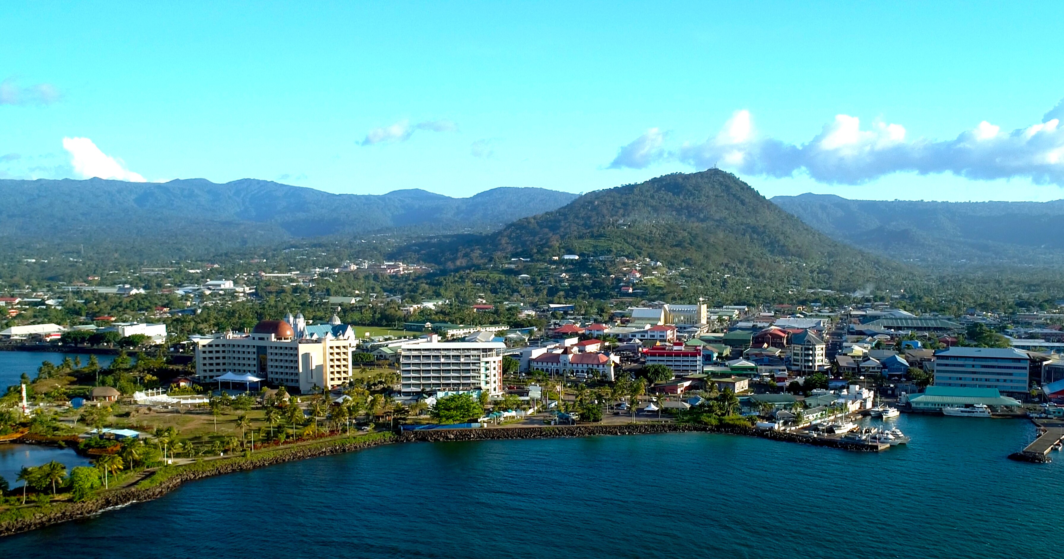 An aerial photo of Apia, Samoa. Mountain ranges in the distance stand above a small city on the waterfront.