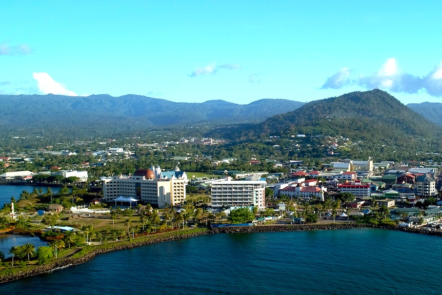 An aerial photo of Apia, Samoa. Mountain ranges in the distance stand above a small city on the waterfront.
