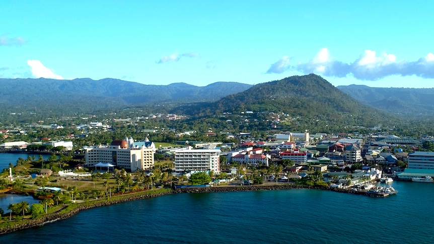 An aerial photo of Apia, Samoa.  Mountain ranges in the distance rise above a small town on the water's edge.