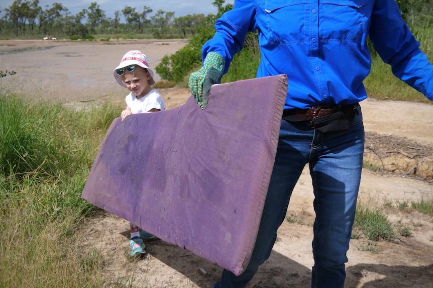 A woman in a blue shirt and a child carry an old mattress near a beach area.