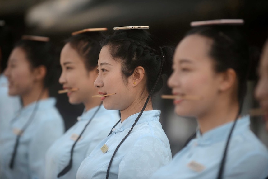 Girls standing in a row practise smiling with mobile phones balanced on their head.