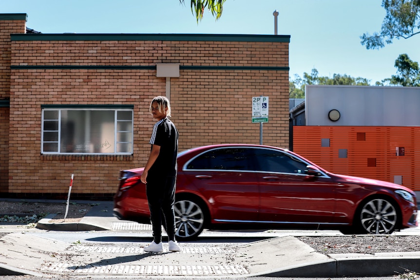 Young man with dark skin and blonder-dyed dreadlocks stands in middle of street with brick wall and car passing