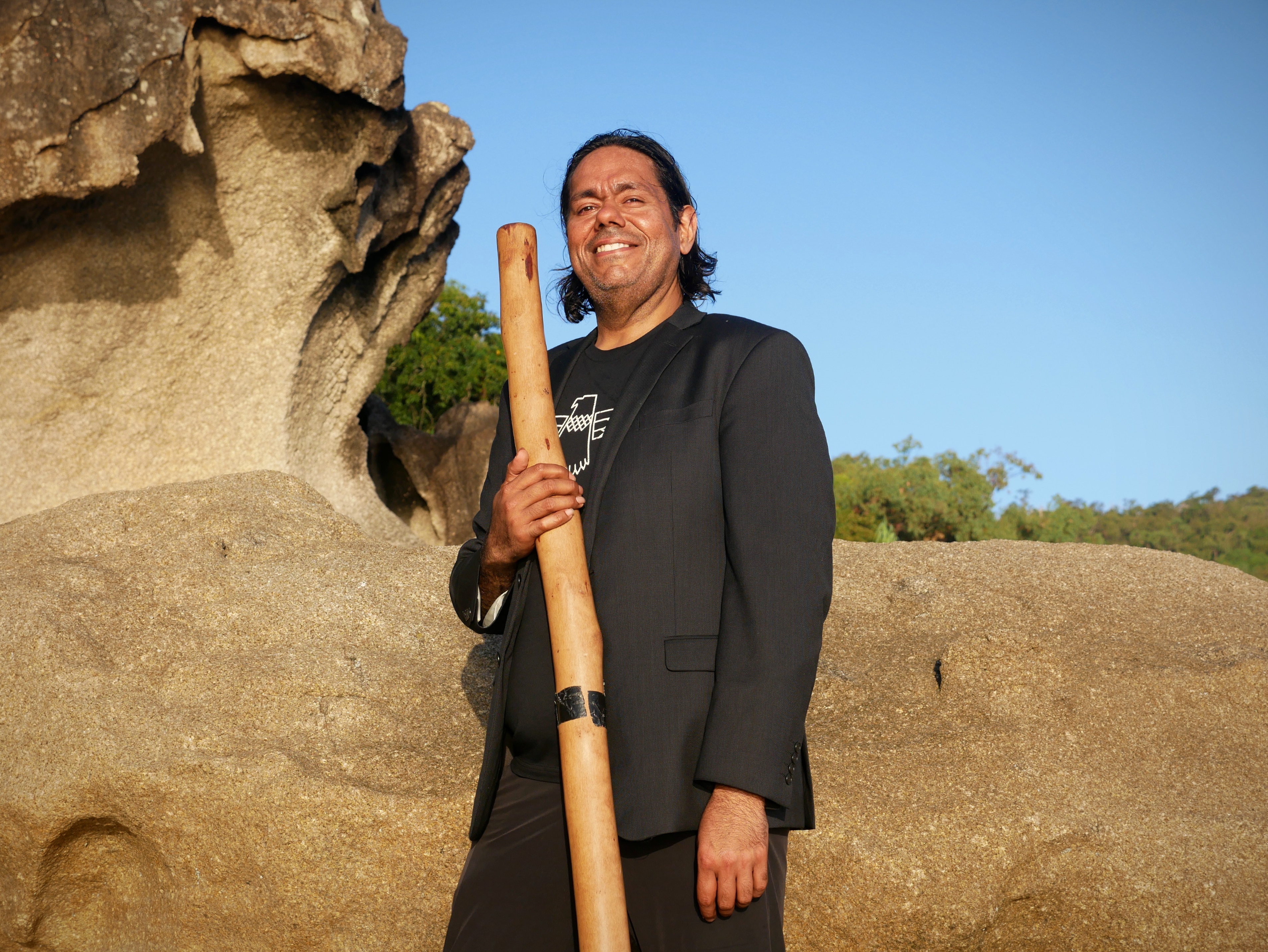 A man stands in front of rocks holding a didgeridoo