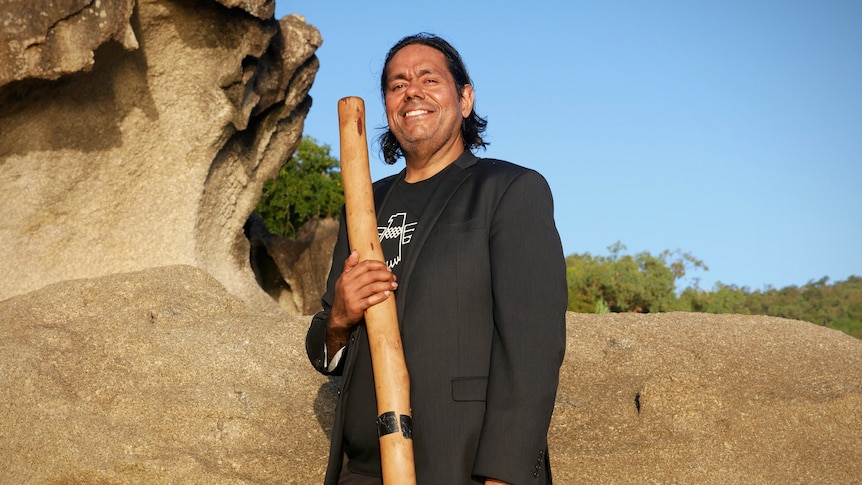 A man stands in front of some rocks holding a didjeridu.
