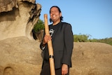 A man stands in front of rocks holding a didgeridoo