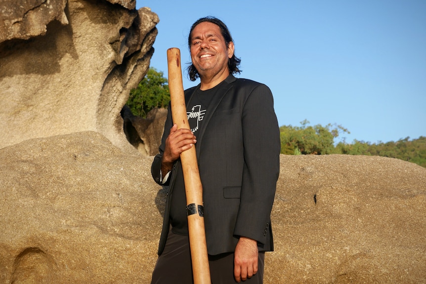 A man stands in front of rocks holding a didgeridoo