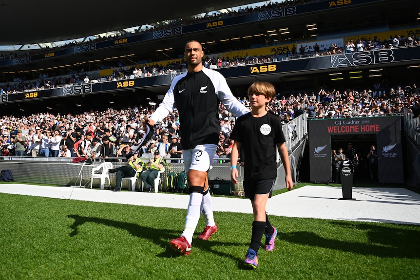 A male soccer player wearing black and white walks out into a stadium alongside a mascot