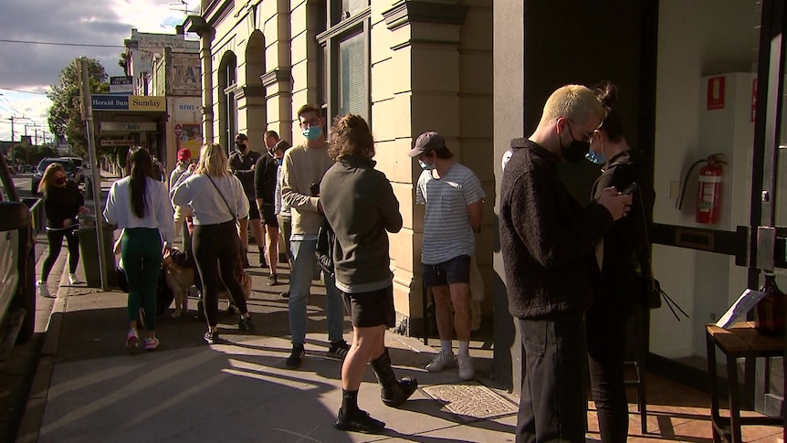 People line up for takeaway drinks outside a pub on a sunny day.