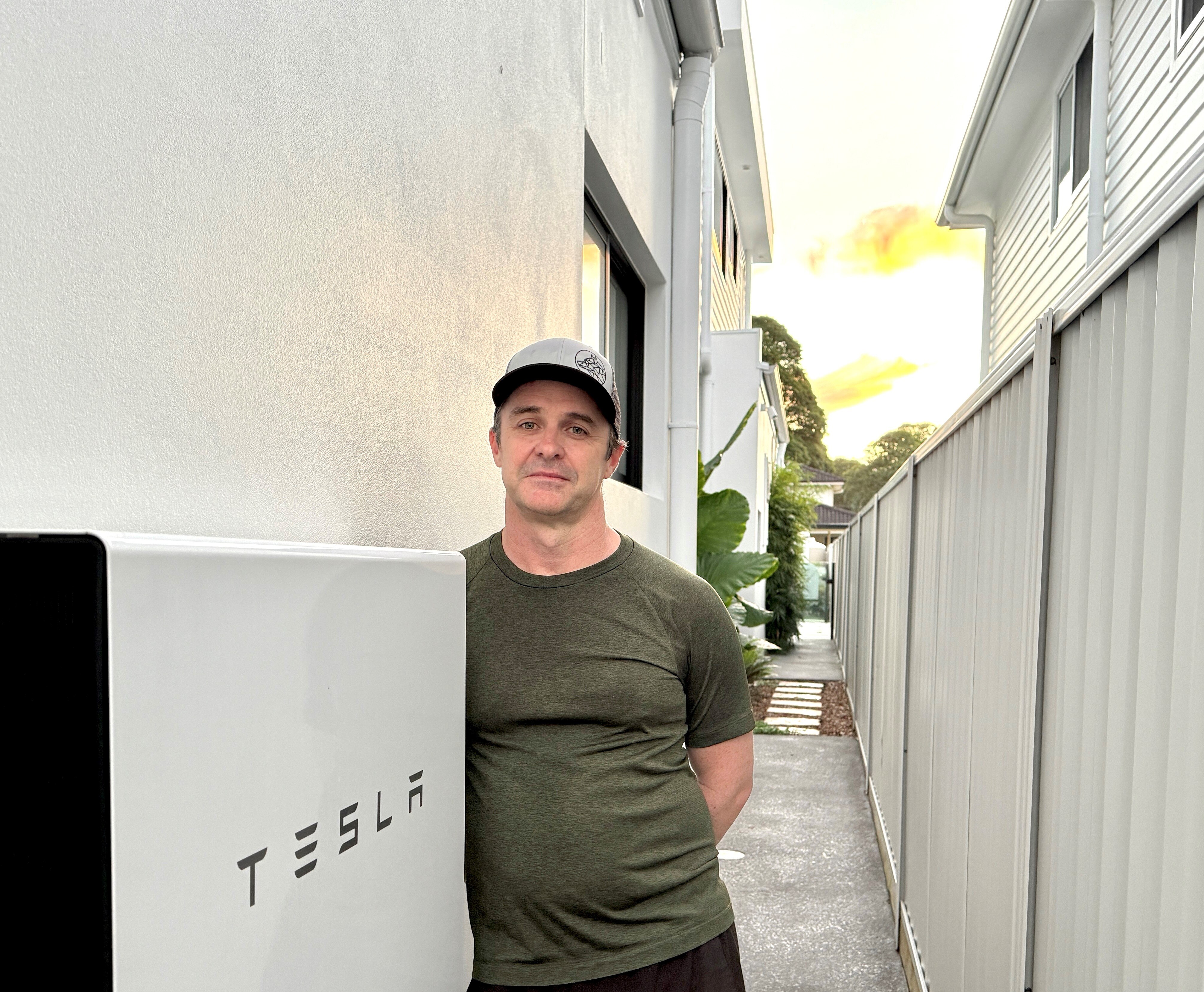 Man wearing baseball cap and khaki green t-shirt standing next to household Tesla battery