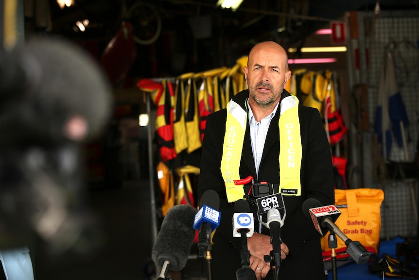 A bald man wearing a yellow life jacket speaks to media at a press conference