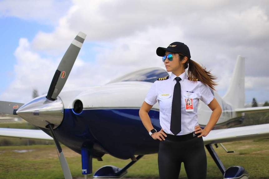 A woman wearing reflective aviators stands with her hands on her hips in front of a plane.