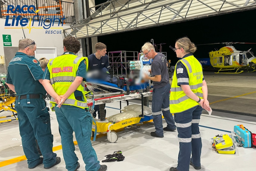 Paramedics stand around a bed in a hangar. A helicopter is visible in the background.