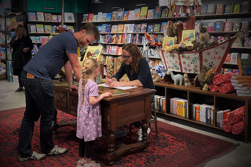A man and his daughter lean against a desk where a man in glasses signs a book.