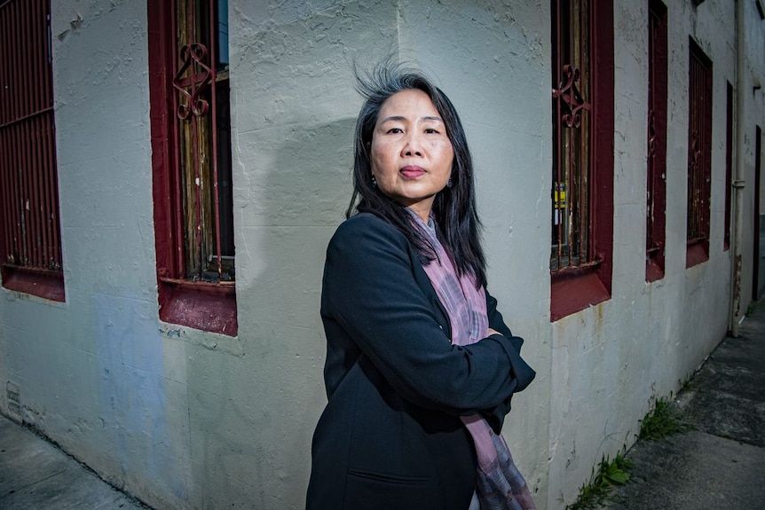 A woman with long dark hair leans against a white wall outside a building.