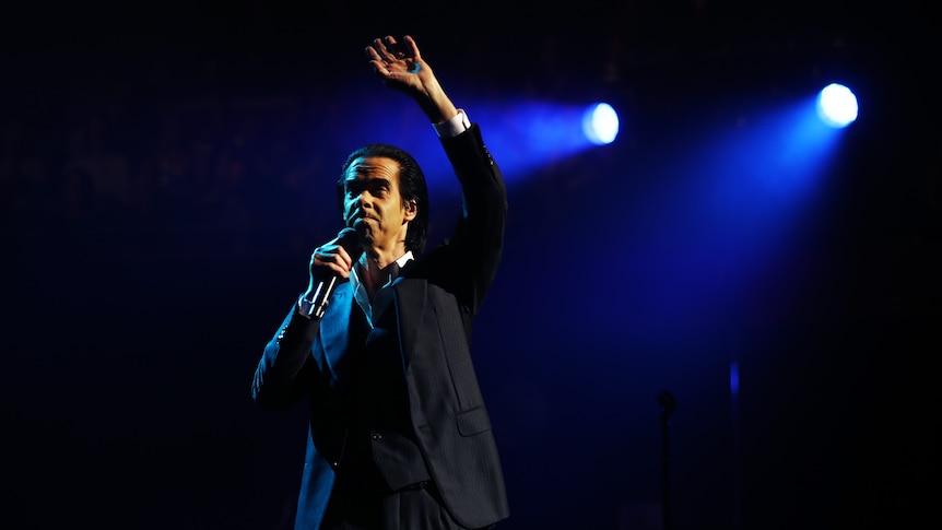 Nick Cave raises his hand above his head while he holds the microphone stand