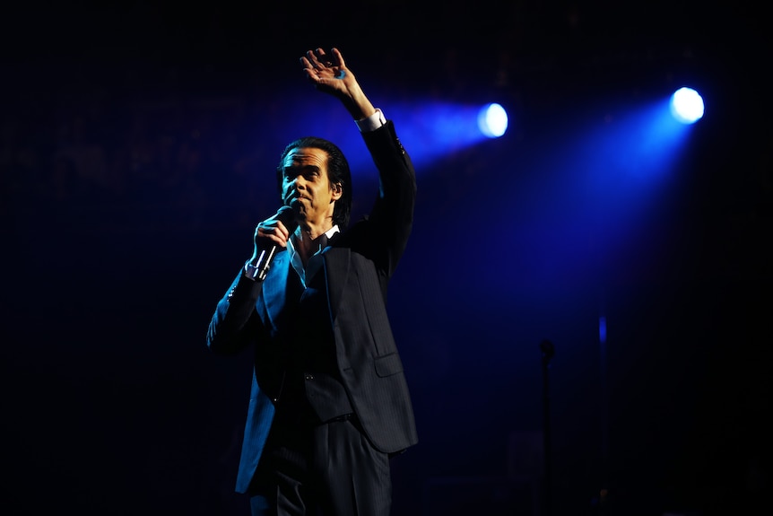 Nick Cave raises his hand above his head while he holds the microphone stand