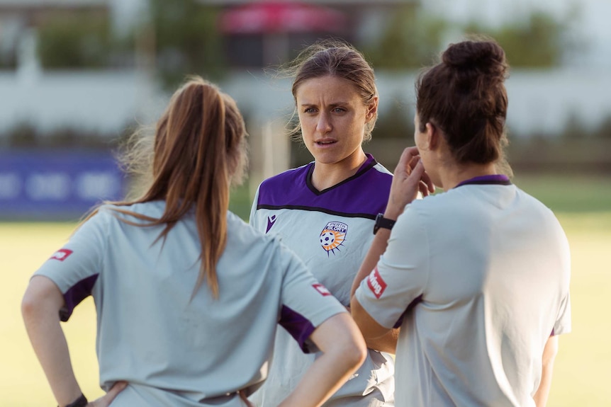 A woman stands and chats with two other girls on a sports field