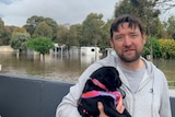 A man standing in front of a flooded caravan park holds his small black dog