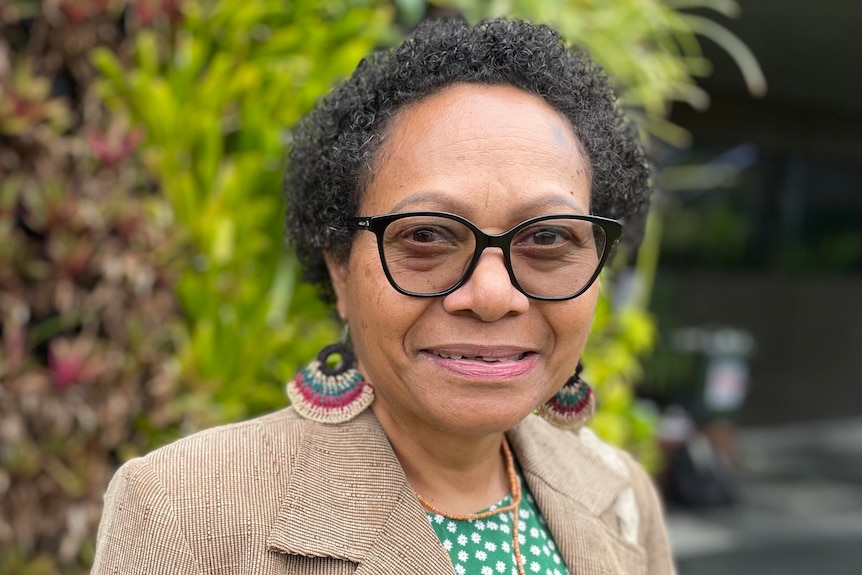 A woman with short dark curly hair wearing colourful woven earrings and glasses smiling in front of a wall of green plants