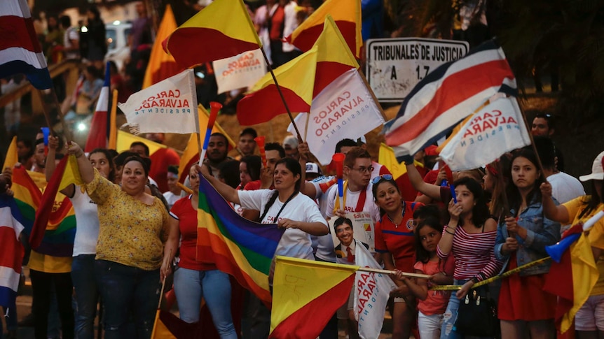 A crowd of people is seen waving rainbow flags and Carlos Alvarado banners.