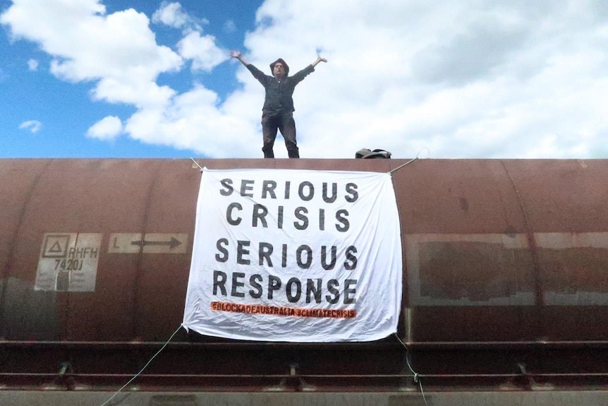 a man stands on a coal wagon with arms outstretched and a sign on wagon saying serious crisis, serious response