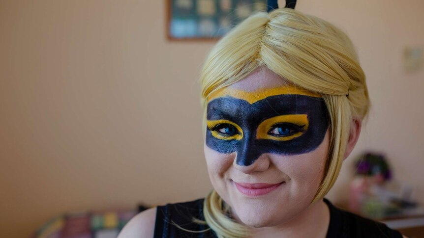 A close up of a woman with a black mask painted on and a blond wig. She smiles at the camera.