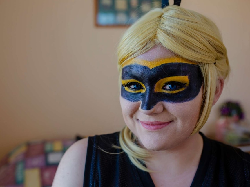 A close up of a woman with a black mask painted on and a blond wig. She smiles at the camera.