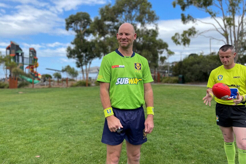 A man wearing an aussie rules umpire's uniform stands on a sports ground and smiles