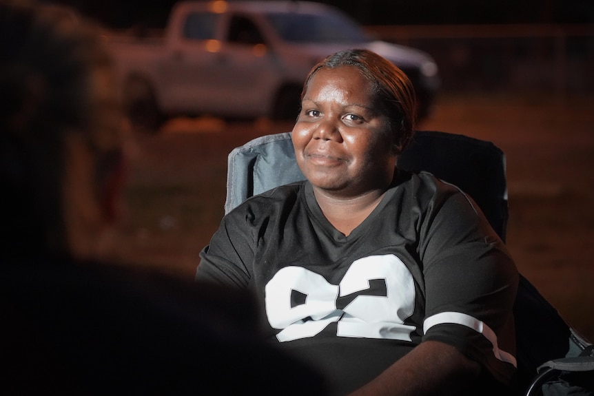 Woman sits on a camping chair at dusk and smiles ahead with a light shining on her face