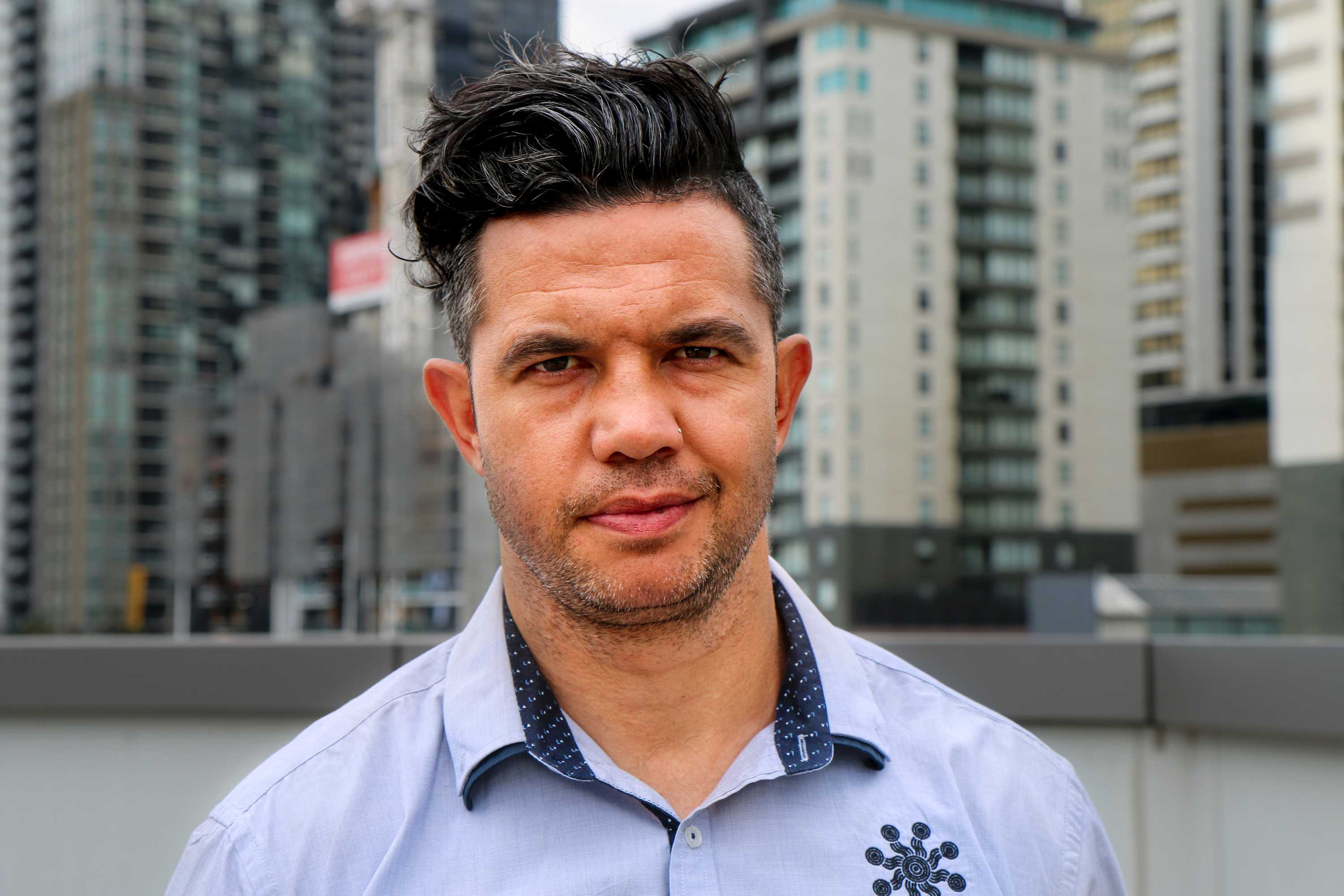 Joe Williams, with black hair and wearing a blue shirt, stands in front of a city background.