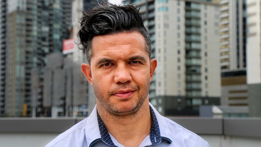 Joe Williams, with black hair and wearing a blue shirt, stands in front of a city background.